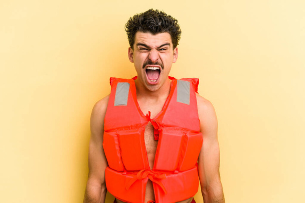 Young caucasian man wearing life jacket isolated on yellow background screaming very angry and aggressive. - Foto, imagen