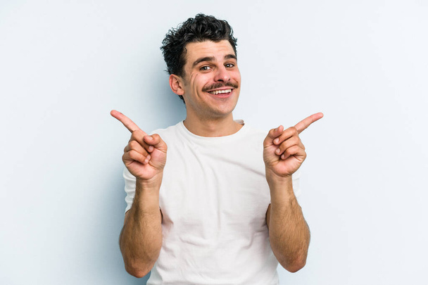 Young caucasian man isolated on blue background pointing to different copy spaces, choosing one of them, showing with finger. - Photo, Image
