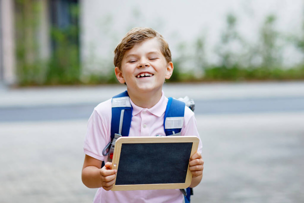 Счастливый маленький мальчик с рюкзаком или сумкой. Школьник по дороге в школу. Здоровый очаровательный ребенок на свежем воздухе на столе В последний день второй класс на немецком языке. Конец школам.
. - Фото, изображение