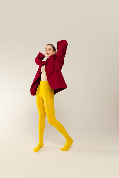 Portrait of young stylish girl in yellow tights and red jacket posing isolated over grey studio background. Bright outfit. Concept of retro fashion, art photography, style, queer, beauty - Фото, изображение