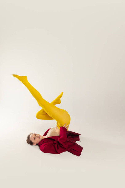 Portrait of young stylish girl in yellow tights and red jacket lying on floor with legs up, posing isolated over grey studio background. Concept of retro fashion, art photography, style, queer, beauty - 写真・画像