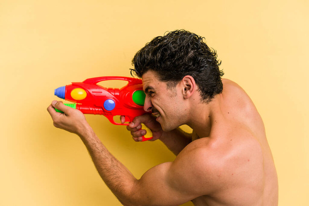 Young caucasian man playing with a water gun isolated on yellow background - Foto, immagini