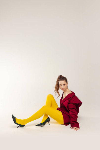 Portrait of stylish young girl sitting, posing in yellow tights, red jacket and heeled shoes isolated over grey studio background. Concept of retro fashion, art photography, style, queer, beauty - Fotó, kép