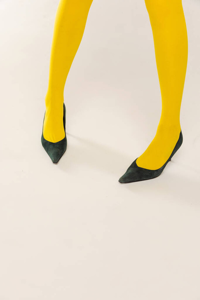 Imagem recortada de pernas femininas em meias amarelas brilhantes e saltos isolados sobre fundo estúdio cinza. Roupa colorida. Conceito de moda retro, fotografia de arte, estilo, queer, beleza - Foto, Imagem