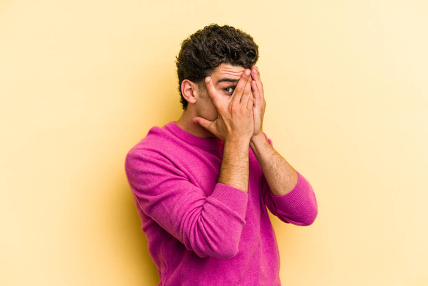 Young caucasian man isolated on yellow background blink through fingers frightened and nervous. - Photo, Image