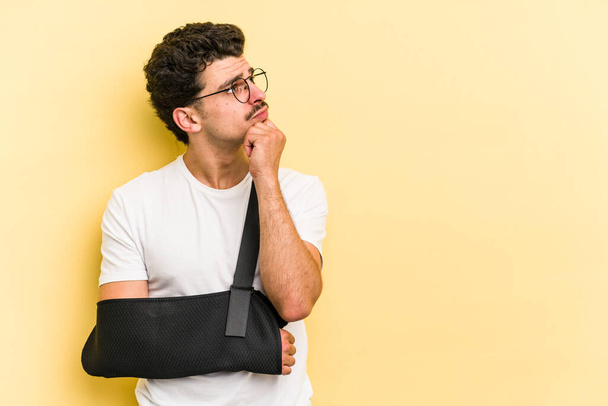 Young caucasian man with broke hand isolated on yellow background looking sideways with doubtful and skeptical expression. - Fotografie, Obrázek