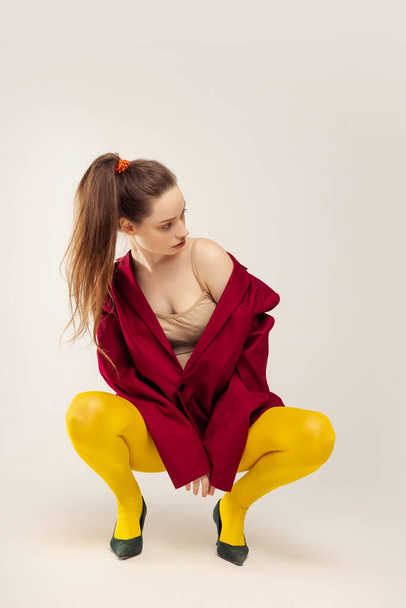 Portret van stijlvol jong meisje in gele maillot, rode jas en hielschoenen, zittend op spijkers, poserend geïsoleerd over grijze achtergrond. Concept van retro mode, kunst fotografie, stijl, queer, schoonheid - Foto, afbeelding