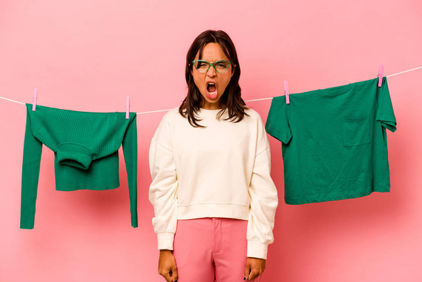 Young hispanic woman holding laundry basket isolated on pink background screaming very angry and aggressive. - 写真・画像