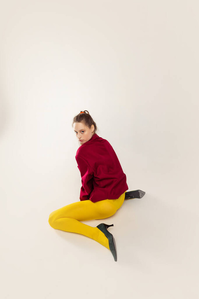 Portrait of young stylish girl in yellow tights, red jacket and heel shoes, sitting, posing isolated over grey studio background. Concept of retro fashion, art photography, style, queer, beauty - Foto, Bild