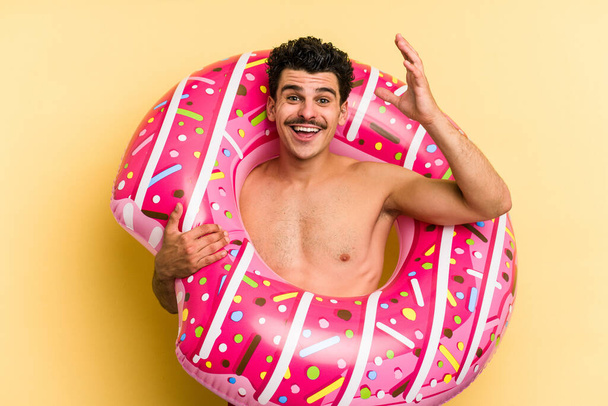 Young caucasian man holding an inflatable donut isolated on yellow background receiving a pleasant surprise, excited and raising hands. - Fotó, kép