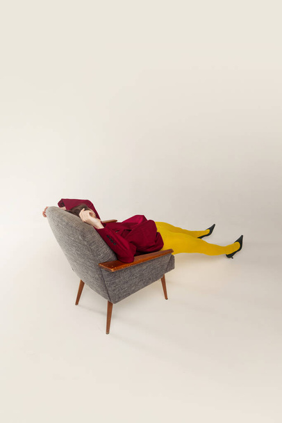 Retrato de niña en medias amarillas y chaqueta roja posando con silla aislada sobre fondo gris estudio. Acostado. Concepto de moda retro, fotografía de arte, estilo, queer, belleza - Foto, Imagen