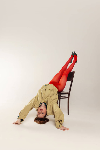 Portrait of young girl in red tights and stylish jacket posing, falling down the chair isolated over grey studio background. Concept of retro fashion, art photography, style, queer, beauty - Foto, afbeelding