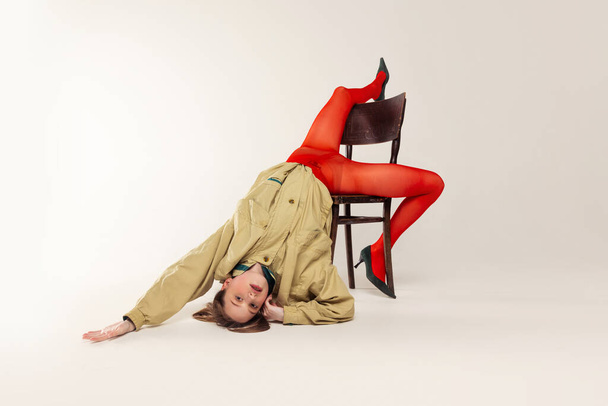 Portret van een jong meisje in rode panty en stijlvolle jas poseren, vallen van de stoel geïsoleerd over grijze studio achtergrond. Concept van retro mode, kunst fotografie, stijl, queer, schoonheid - Foto, afbeelding