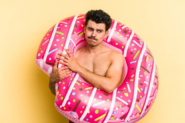 Young caucasian man holding an inflatable donut isolated on yellow background confused, feels doubtful and unsure. - Foto, Imagen