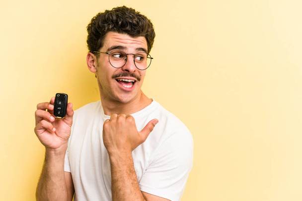 Young caucasian man holding car keys isolated on yellow background points with thumb finger away, laughing and carefree. - Foto, Imagen