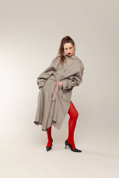 Portrait of young stylish girl posing in red tights and coat posing isolated over grey studio background. Blushing cheeks. Concept of retro fashion, art photography, style, queer, beauty - 写真・画像