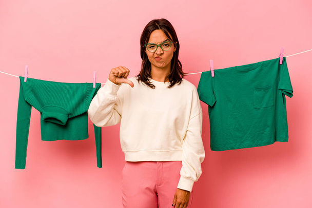 Young hispanic woman holding laundry basket isolated on pink background showing a dislike gesture, thumbs down. Disagreement concept. - Photo, Image