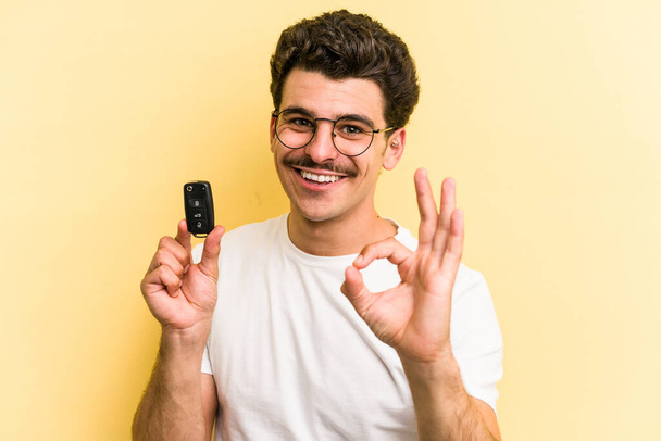 Young caucasian man holding car keys isolated on yellow background cheerful and confident showing ok gesture. - 写真・画像
