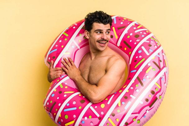 Young caucasian man holding an inflatable donut isolated on yellow background looks aside smiling, cheerful and pleasant. - Valokuva, kuva