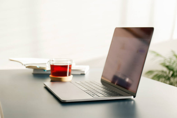 Home office concept. Designated work from home area near the window. Modern laptop with blurred screen and glass cup of tea on table. Close up, copy space, interior background. - Valokuva, kuva