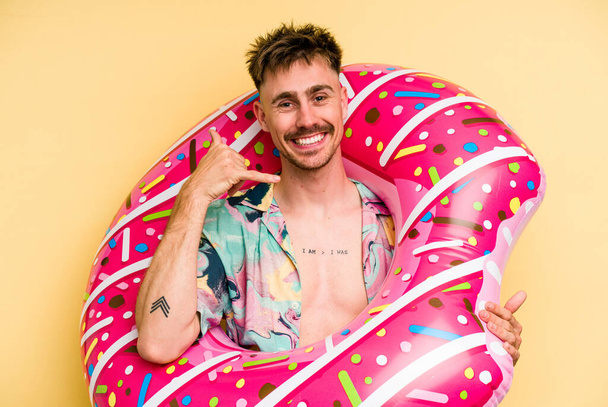 Young caucasian man holding an inflatable donut isolated on yellow background showing a mobile phone call gesture with fingers. - Foto, Bild