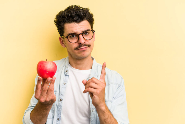 Young caucasian man holding an apple isolated on yellow background showing number one with finger. - Fotografie, Obrázek