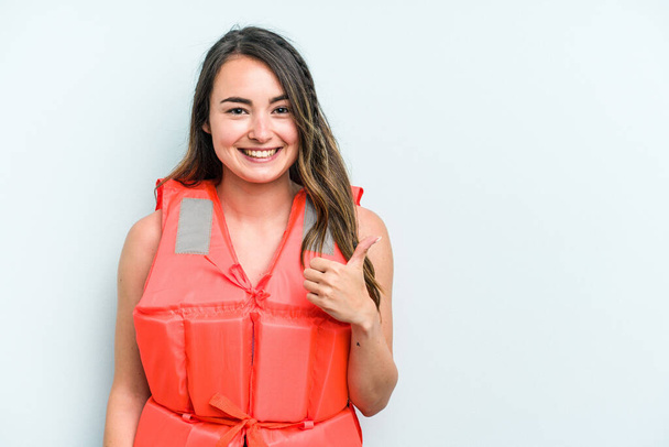 Young caucasian woman wearing life jacket isolated on blue background smiling and raising thumb up - 写真・画像