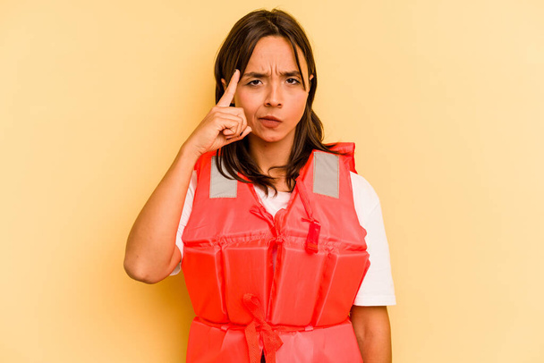 Молода іспаномовна жінка тримає рятувальний жилет ізольовано на жовтому тлі, вказуючи храм пальцем, думаючи, зосереджений на завданні
. - Фото, зображення