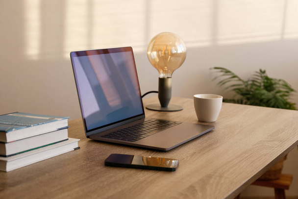 Blank screen laptop, cell phone and a stack of books on wooden table. Close up, copy space, white wall background. - 写真・画像