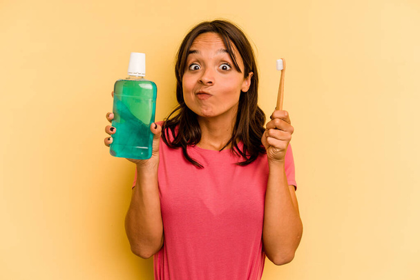 Young hispanic woman holding mouthwash isolated on yellow background - 写真・画像