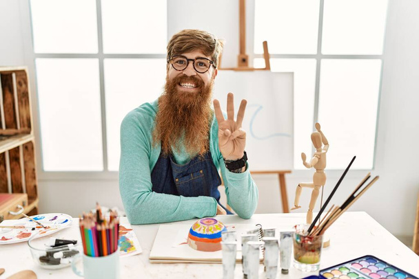 Rudowłosy mężczyzna z długą brodą malujący glinianą miskę w studiu sztuki pokazujący i wskazujący palcami numer trzy uśmiechając się pewny siebie i szczęśliwy.  - Zdjęcie, obraz