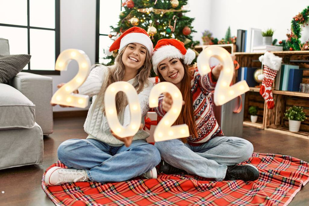 Женская пара празднует Новый 2022 год сидя на полу у рождественской елки дома - Фото, изображение
