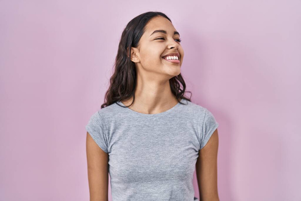 若いブラジルの女性の顔に笑顔で横に離れて見てピンクの背景にカジュアルなTシャツを着て、自然な表情。自信を持って.  - 写真・画像