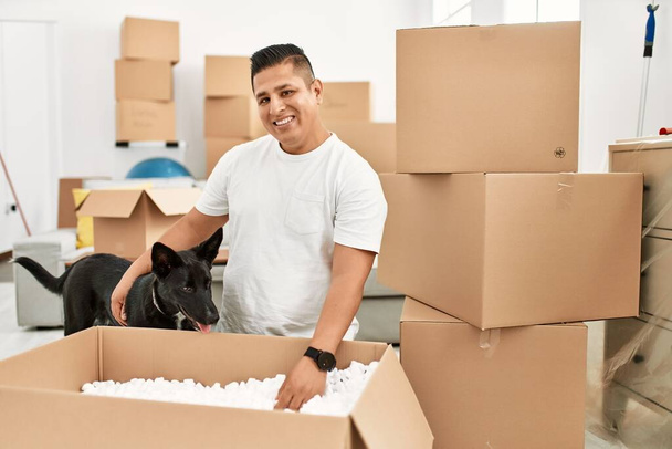 Jeune homme latino souriant heureux boîte de carton de déballage à la nouvelle maison avec chien. - Photo, image