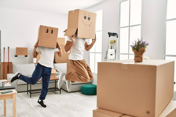 Two hispanic men couple playing with funny cardboard boxes on head at new home - Photo, Image