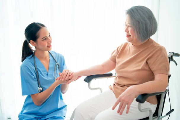 Asiatische Krankenschwester oder Arzt sitzen auf dem Boden und halten die Hände der Seniorin sitzen vor Vorhang im Wohnzimmer. - Foto, Bild