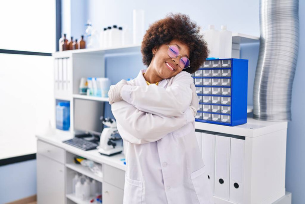 Νεαρή Αφροαμερικάνα που δουλεύει σε επιστημονικό εργαστήριο αγκαλιάζοντας τον εαυτό της χαρούμενη και θετική, χαμογελαστή αυτοπεποίθηση. αυτοαγάπη και αυτοφροντίδα  - Φωτογραφία, εικόνα