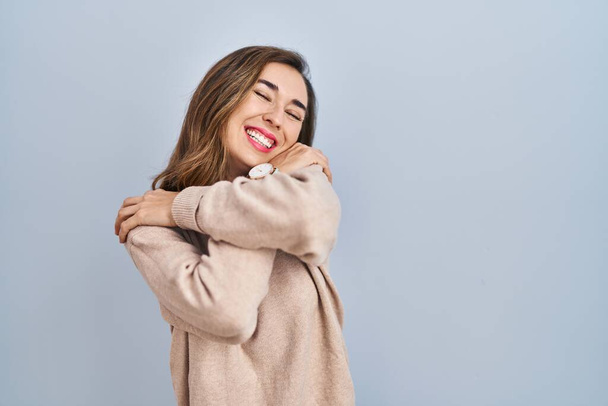 Young woman standing over isolated background hugging oneself happy and positive, smiling confident. self love and self care  - Фото, изображение