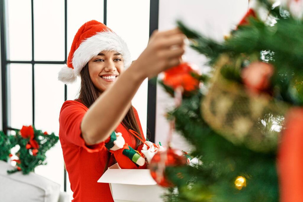 Молода латинка відкриває подарунковий пакет, сидячи на різдвяному декорі вдома
 - Фото, зображення