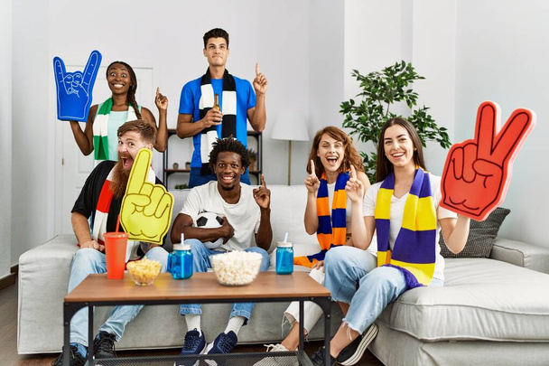 Ομάδα φίλων που υποστηρίζουν την ομάδα ποδοσφαίρου βλέποντας τηλεόραση al σπίτι κάθεται στον καναπέ έκπληκτος με μια ιδέα ή ερώτηση δείχνοντας δάχτυλο με χαρούμενο πρόσωπο, νούμερο ένα  - Φωτογραφία, εικόνα