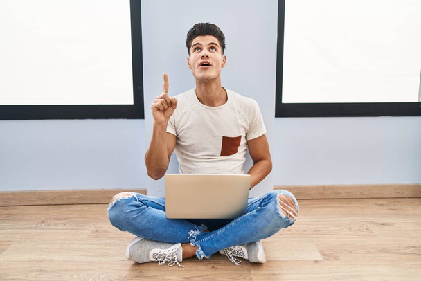 Young hispanic man using laptop at home amazed and surprised looking up and pointing with fingers and raised arms.  - 写真・画像