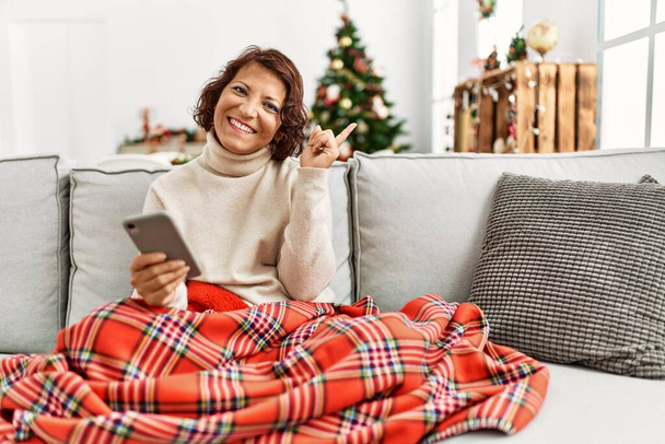 Moyen Age femme hispanique en utilisant smartphone assis sur le canapé par arbre de Noël souriant heureux pointant avec la main et le doigt sur le côté  - Photo, image