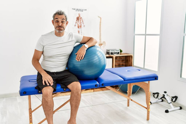 Middle age hispanic man at pain recovery clinic holding pilates ball puffing cheeks with funny face. mouth inflated with air, crazy expression.  - Fotoğraf, Görsel