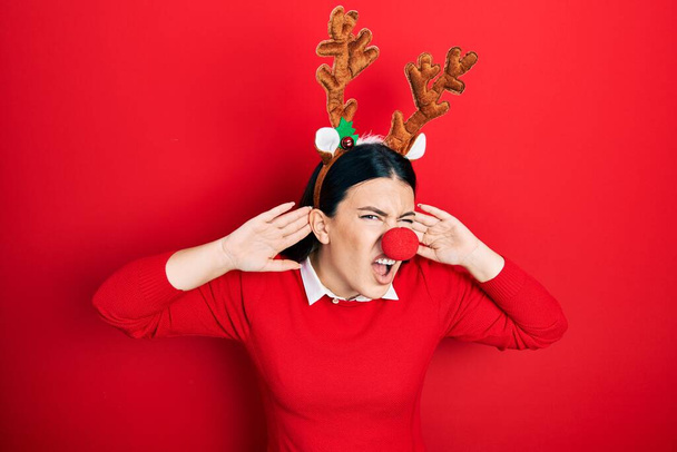 Young hispanic woman wearing deer christmas hat and red nose trying to hear both hands on ear gesture, curious for gossip. hearing problem, deaf  - Φωτογραφία, εικόνα