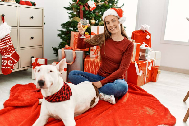 Nuori valkoihoinen nainen juo samppanjaa istuen koiran kanssa joulukuusi kotona - Valokuva, kuva