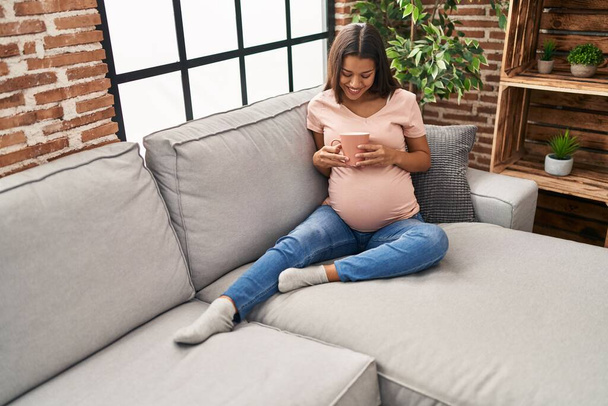 Giovane donna latina incinta che beve caffè seduta sul divano a casa - Foto, immagini