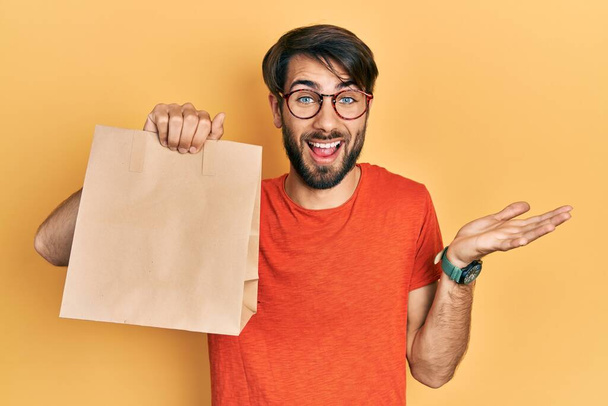Young hispanic man holding paper bag with bread celebrating achievement with happy smile and winner expression with raised hand  - Foto, afbeelding