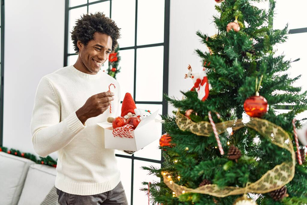 Junger afrikanisch-amerikanischer Mann lächelt glücklich beim Schmücken des Weihnachtsbaums zu Hause. - Foto, Bild