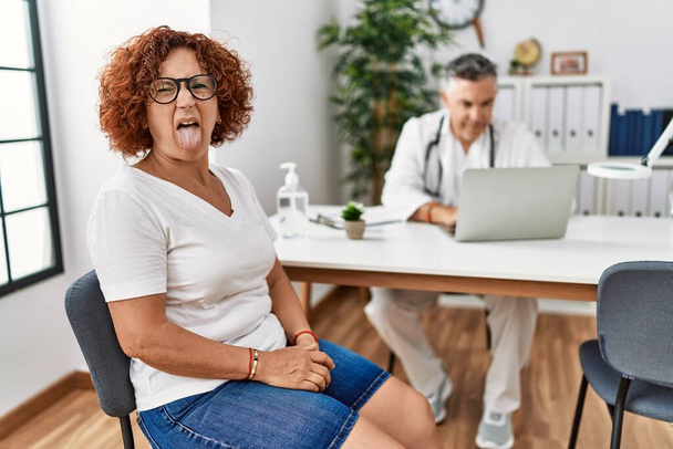 Donna anziana seduta ad un appuntamento dal medico che tira fuori la lingua felice con espressione divertente. concetto di emozione.  - Foto, immagini