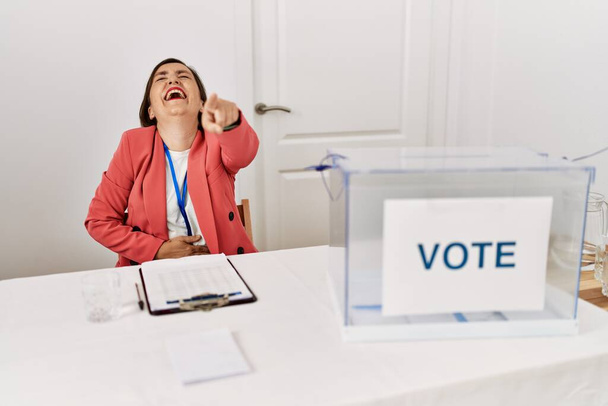 Beautiful middle age hispanic woman at political election sitting by ballot laughing at you, pointing finger to the camera with hand over body, shame expression  - Foto, Imagen